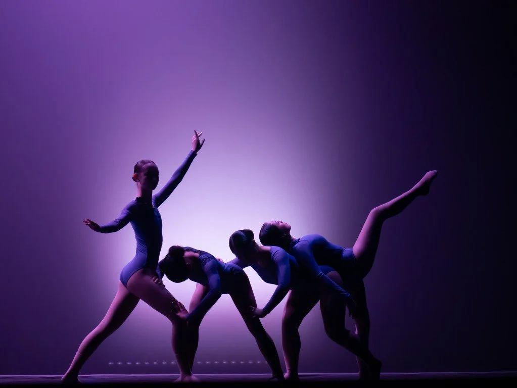 Four ballet dancers performing on stage with purple lighting. The dancer on the left is standing with one arm raised, while the other three are bent forward and holding hands, with one leg extended upward.