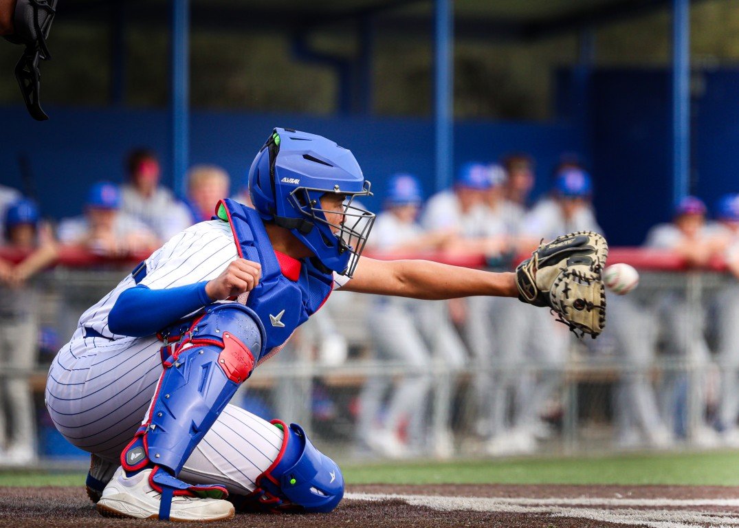 A baseball catcher in a crouched position, wearing a blue helmet, leg guards, and a chest protector, catching a baseball with their glove during a game.