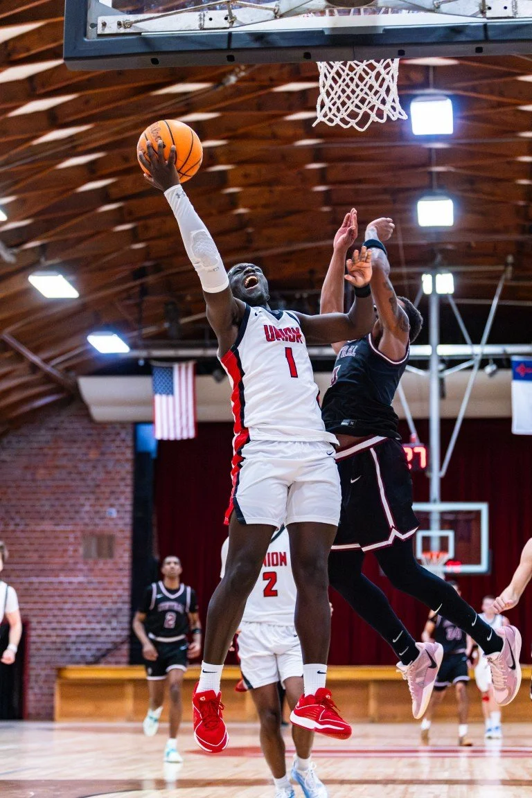 A basketball player in a white uniform jumps to shoot the ball while a defender in black attempts to block the shot inside a gymnasium.