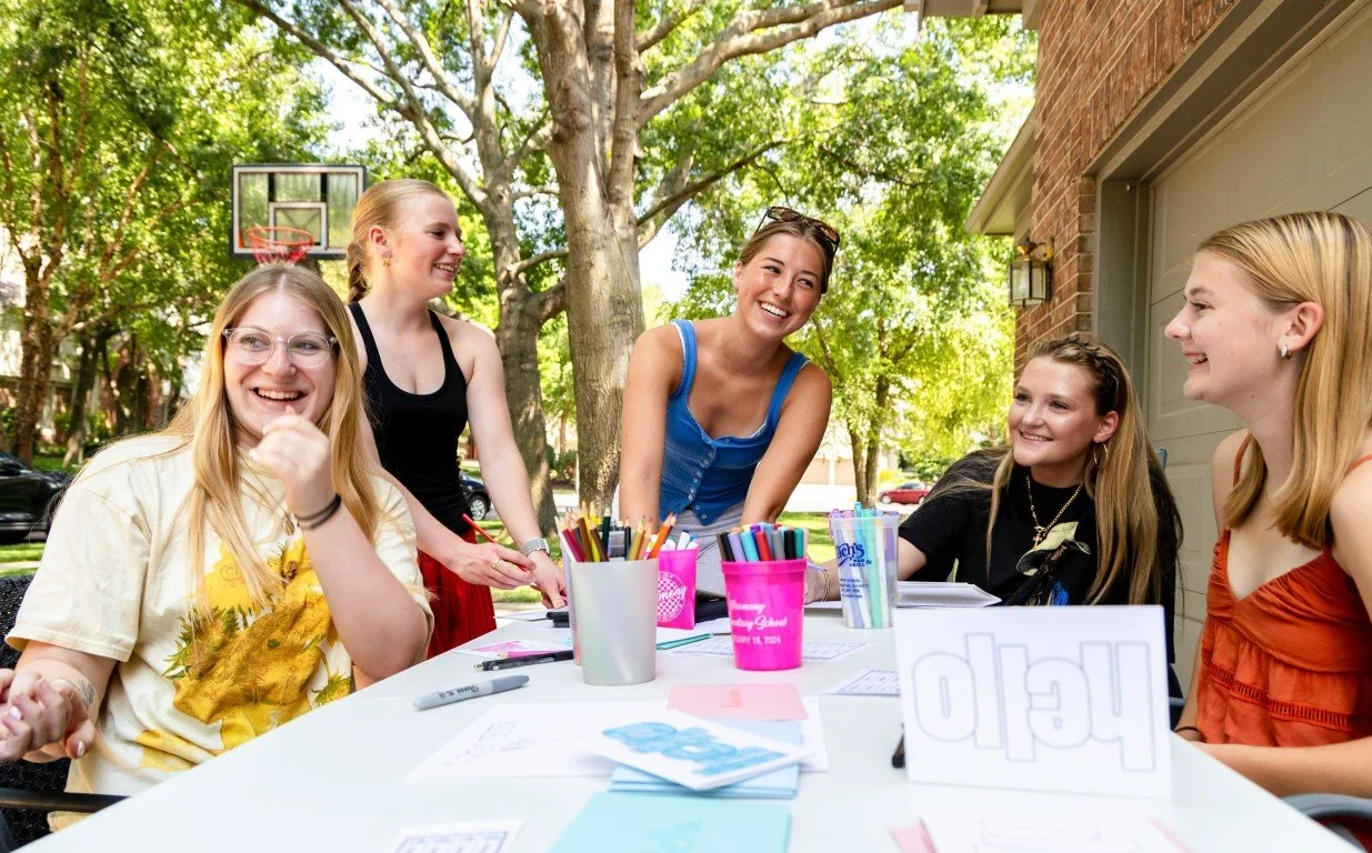 Five young women sitting and standing around a table outside, smiling and laughing, with school supplies and notebooks on the table, trees and a basketball hoop in the background.