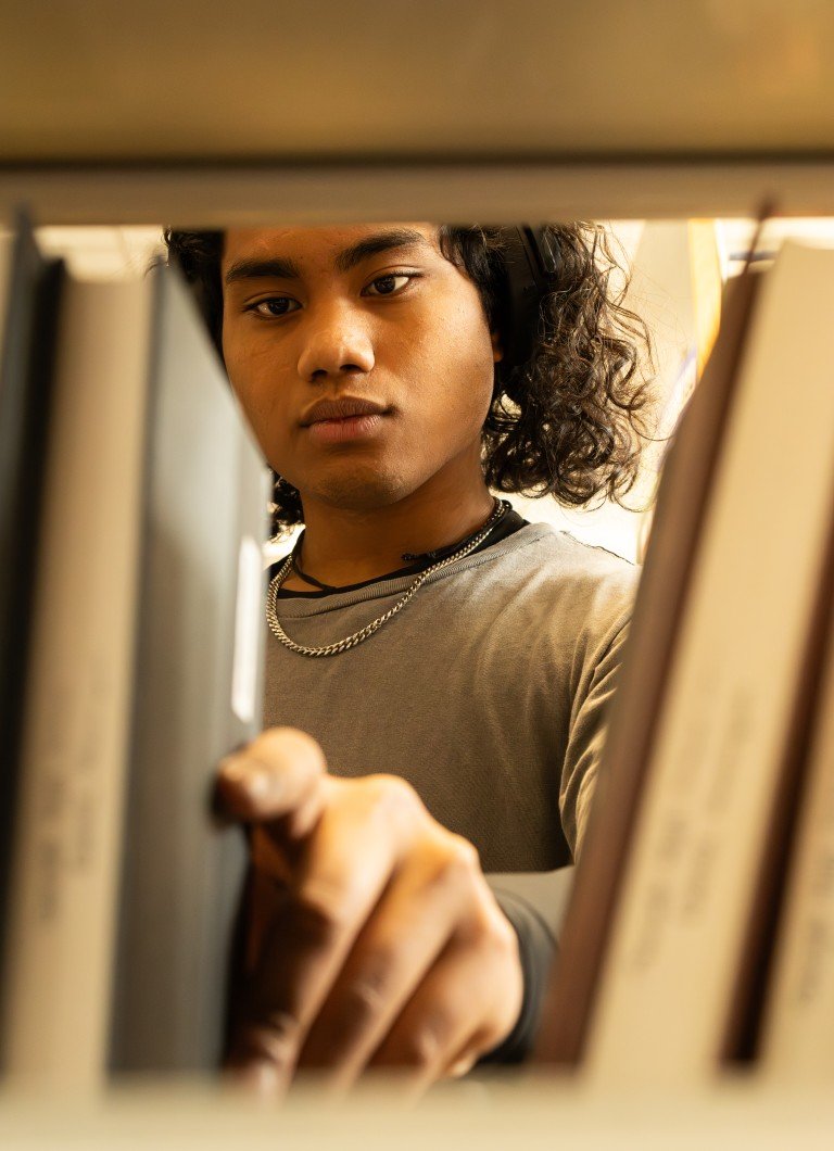 A person with curly hair and a necklace is searching for a book on a bookshelf.