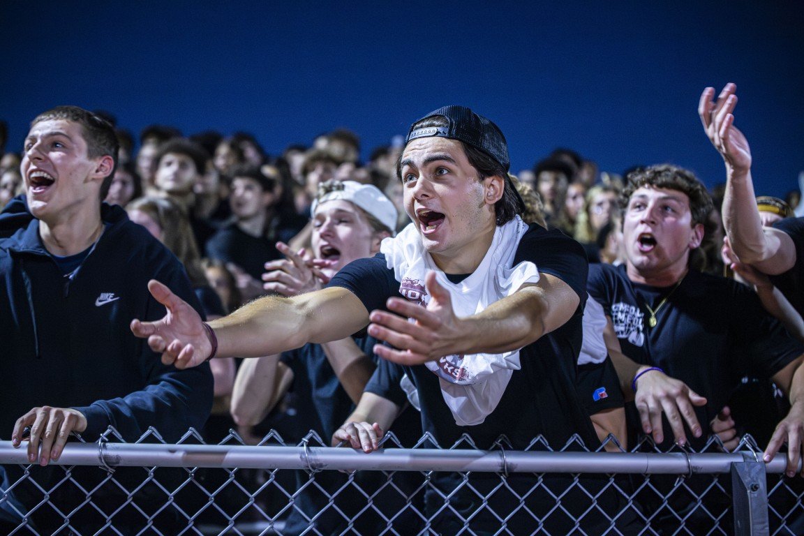 A group of young people, mostly teenage boys, cheering enthusiastically at an outdoor event at night, standing behind a chain link fence.