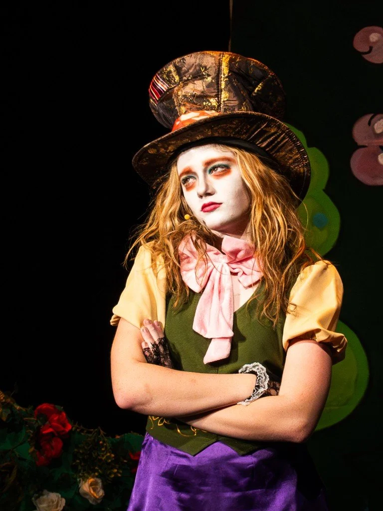 A woman dressed as a whimsical character with a tall, shiny gold and black hat, painted white face, colorful makeup, yellow and pink clothing, a pink bow tie, and purple skirt, standing with arms crossed, in a theatrical setting with green circular d