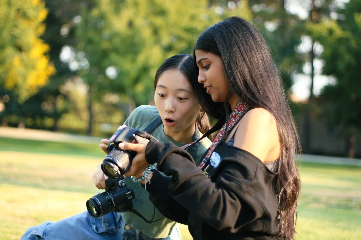 Two young women outdoors, one showing the camera to the other with surprised expressions, surrounded by trees.
