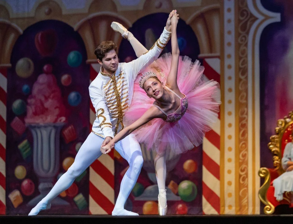Two ballet dancers perform on stage, with the male dancer holding the female dancer's hand while she extends her leg in a ballet pose. Both wear costumes with the female in a pink tutu and the male in a white costume with gold accents, against a colo