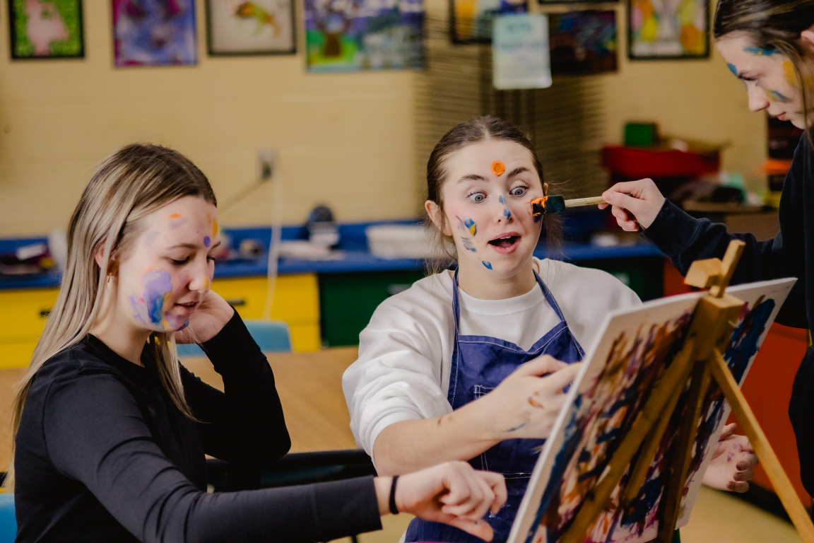 Three girls with colorful paint on their faces creating art in an art classroom with paintings on the wall.