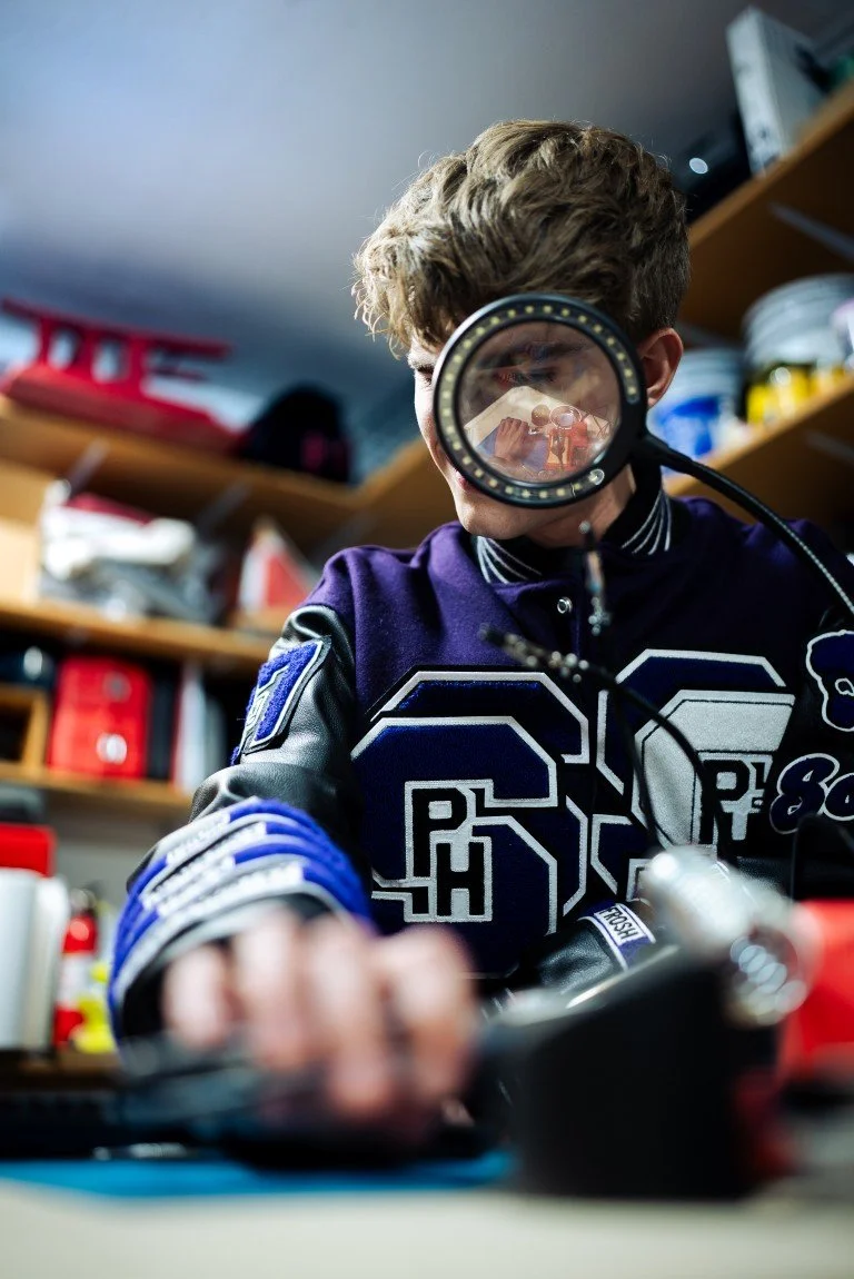 A young person with brown hair is sitting at a workbench, holding a magnifying glass in front of their face, looking through it at an object on the table. The background shows shelves with various tools and supplies.