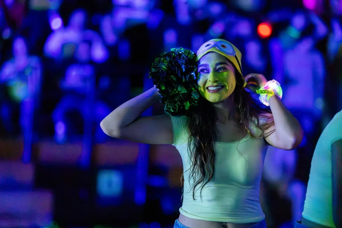 A woman wearing a Minions-themed hat and glow bracelets, smiling and holding her hair back in a lively, colorful party or celebration setting.