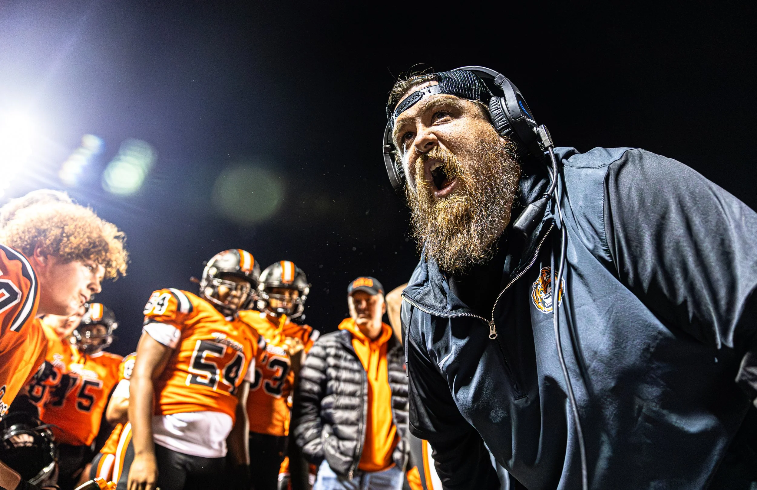 A football coach passionately shouting at his team during a night game, with players in orange uniforms listening attentively.