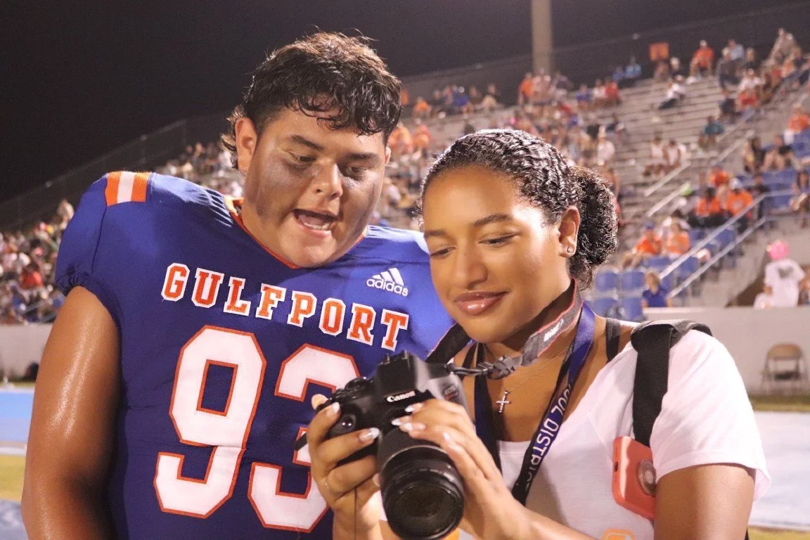 A young male football player in a blue jersey with 'GULFPORT 93' on it stands next to a young woman with a camera and a medal around her neck, at a sports stadium at night with spectators in the background.