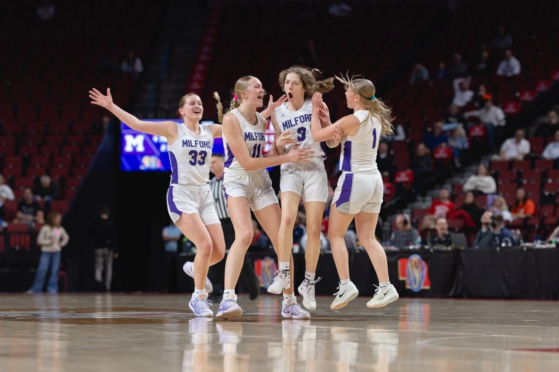 Four female basketball players from Milford team celebrate on the court, with one player jumping in the air, in an arena with spectators in the background.