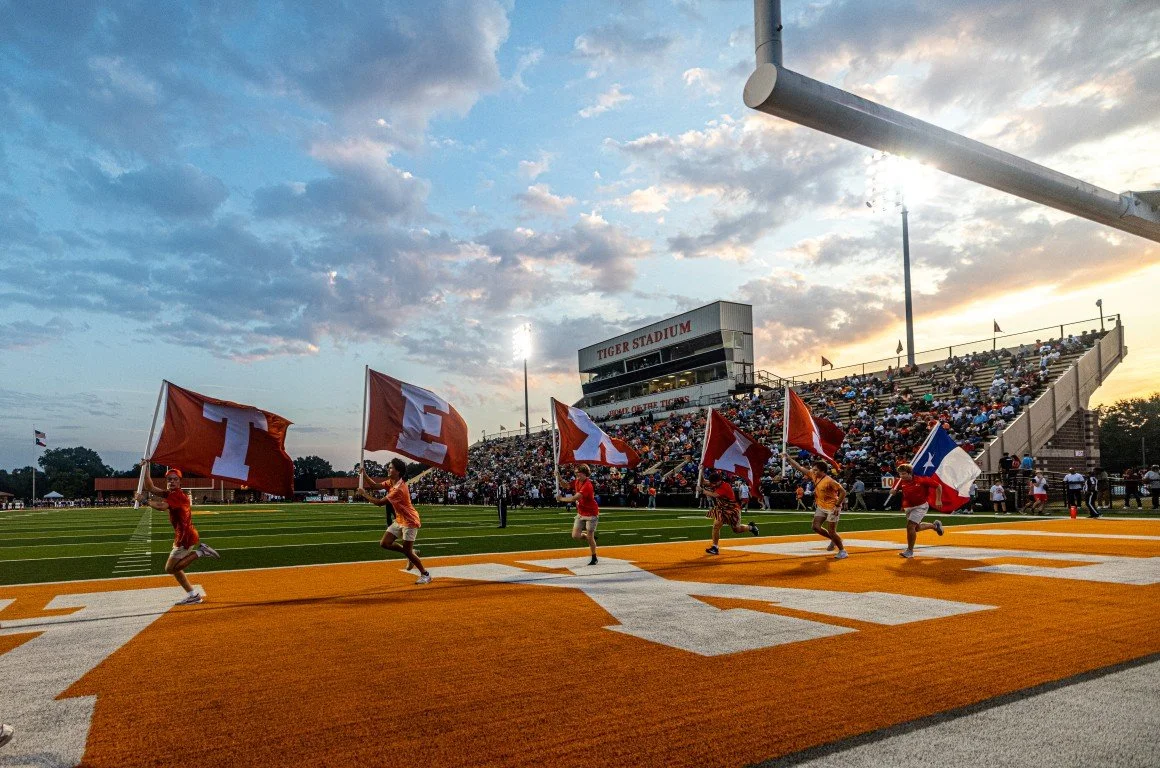 Children holding large flags on a football field at sunset during a game at Tiger Stadium with spectators in the stands.