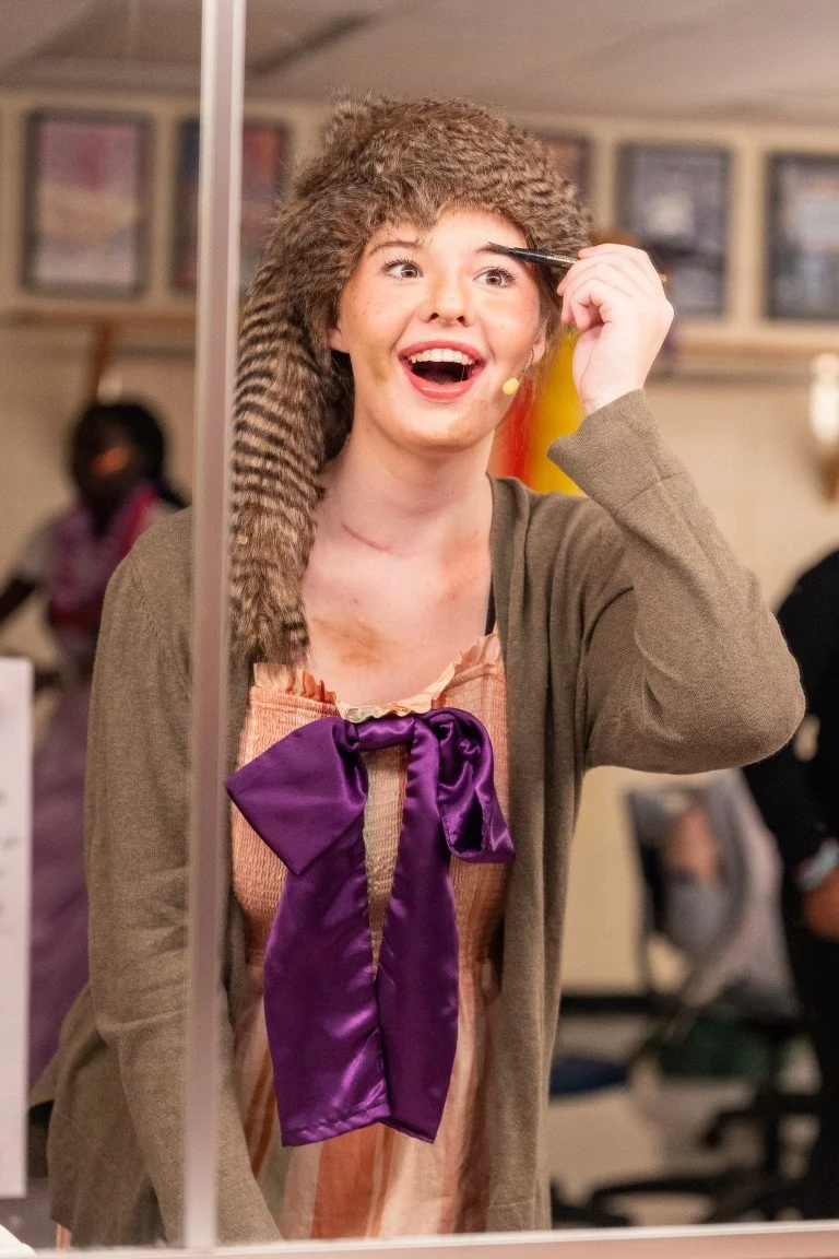 A woman with short hair and makeup is smiling and looking into a mirror, holding a makeup brush to her eyebrow, wearing a bear hat, beige jacket, and a purple bow tie
