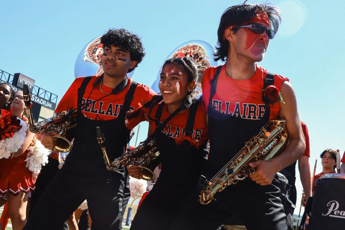 Three young people in red and black band uniforms, holding saxophones, walking together at an outdoor event on a sunny day.