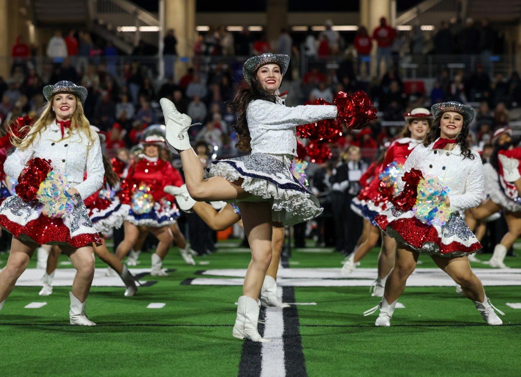 Cheerleaders performing a dance routine on a football field during a game, dressed in white and red costumes with silver hats, holding pom-poms, with spectators in the background.