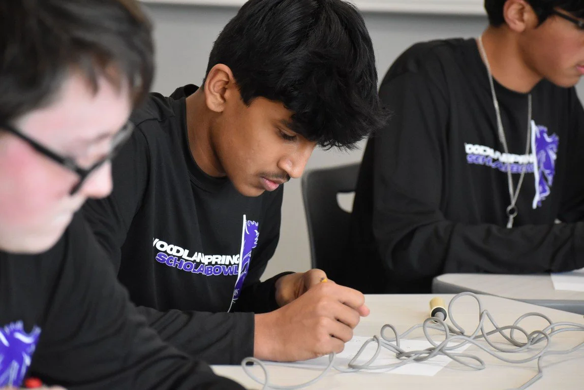 Three students in black shirts working on a science project at a desk with wires and a small cylindrical object.