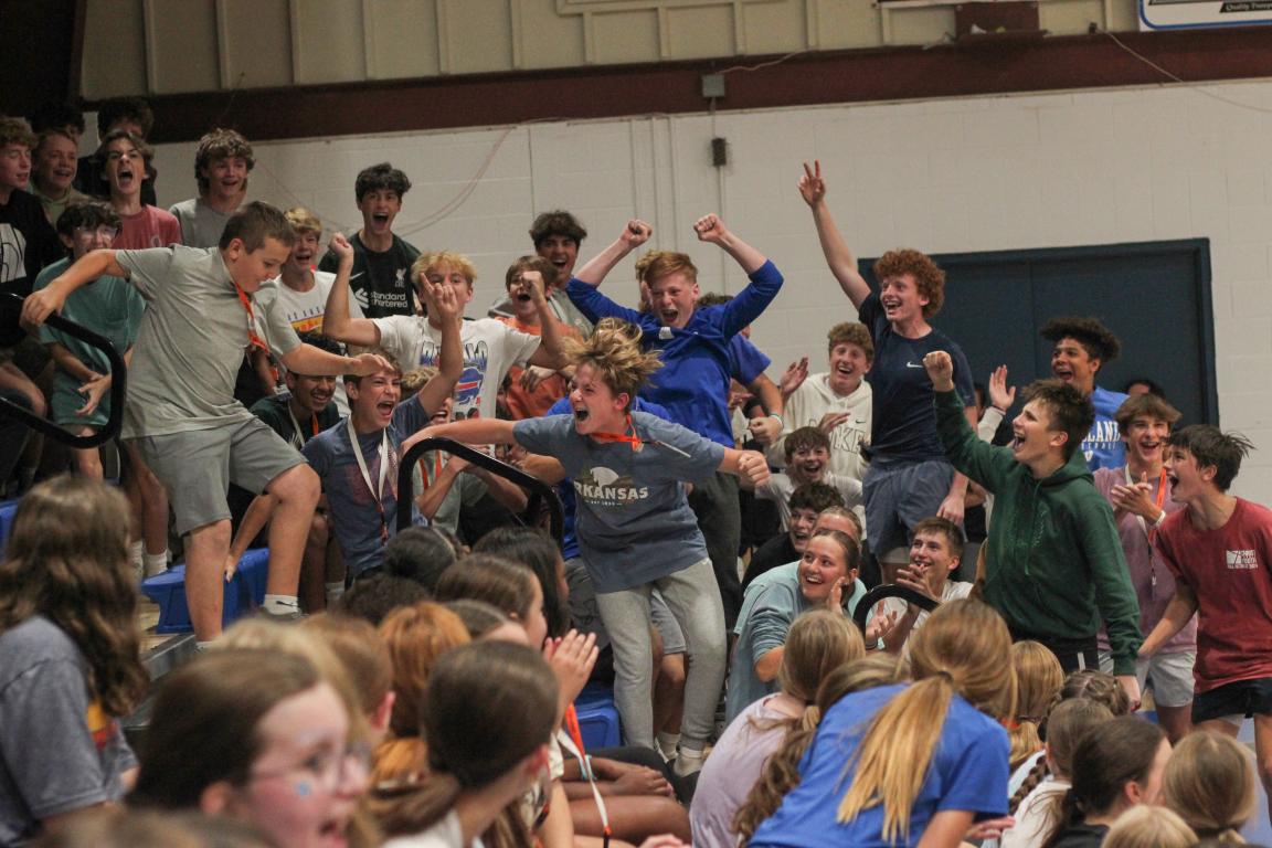 A large group of kids and teenagers celebrating and cheering at an indoor event, some are standing on chairs or tables, many with arms raised or making gestures of excitement.