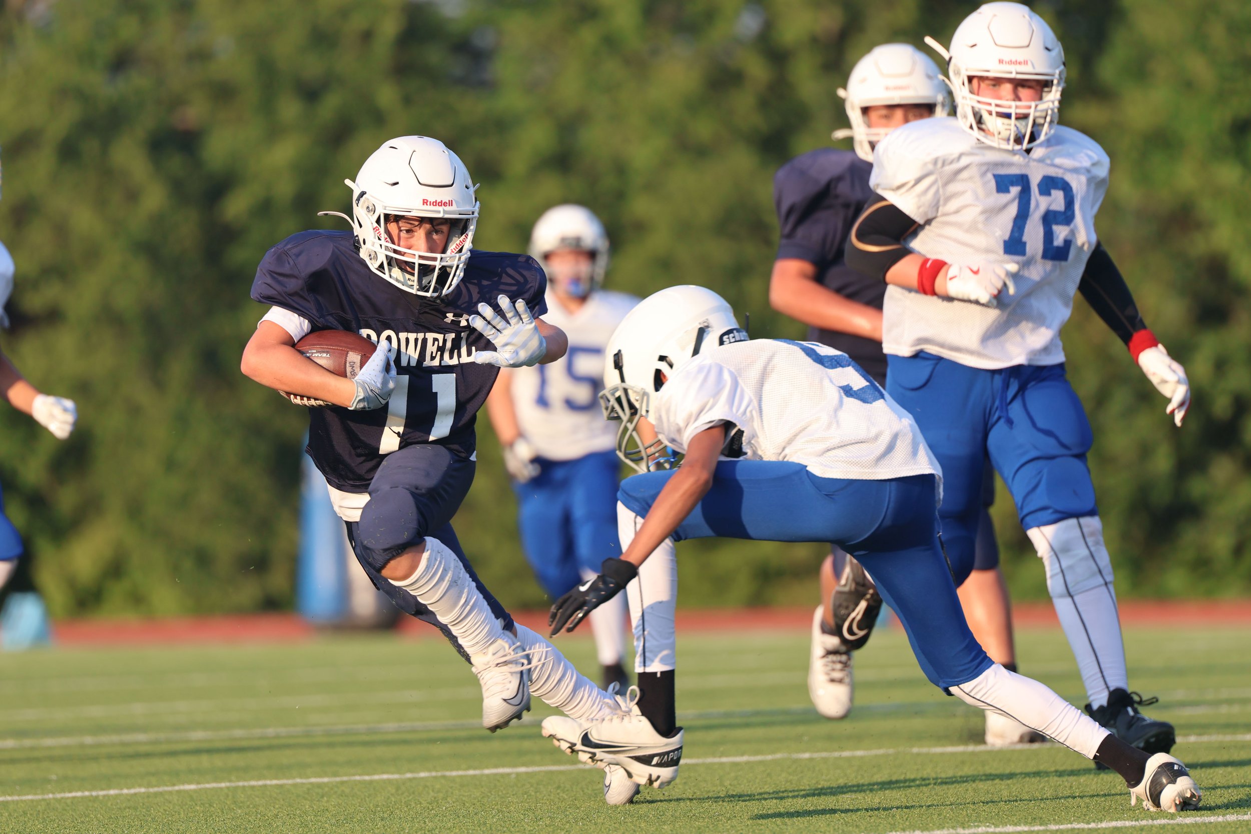 Young football players in action during a game, wearing helmets and uniforms, with a background of green trees.