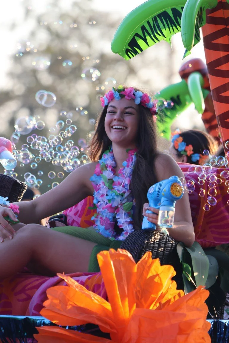 A young woman in a flower lei and a floral headband laughing and having fun, surrounded by bubbles and colorful inflatables during a festive outdoor event.