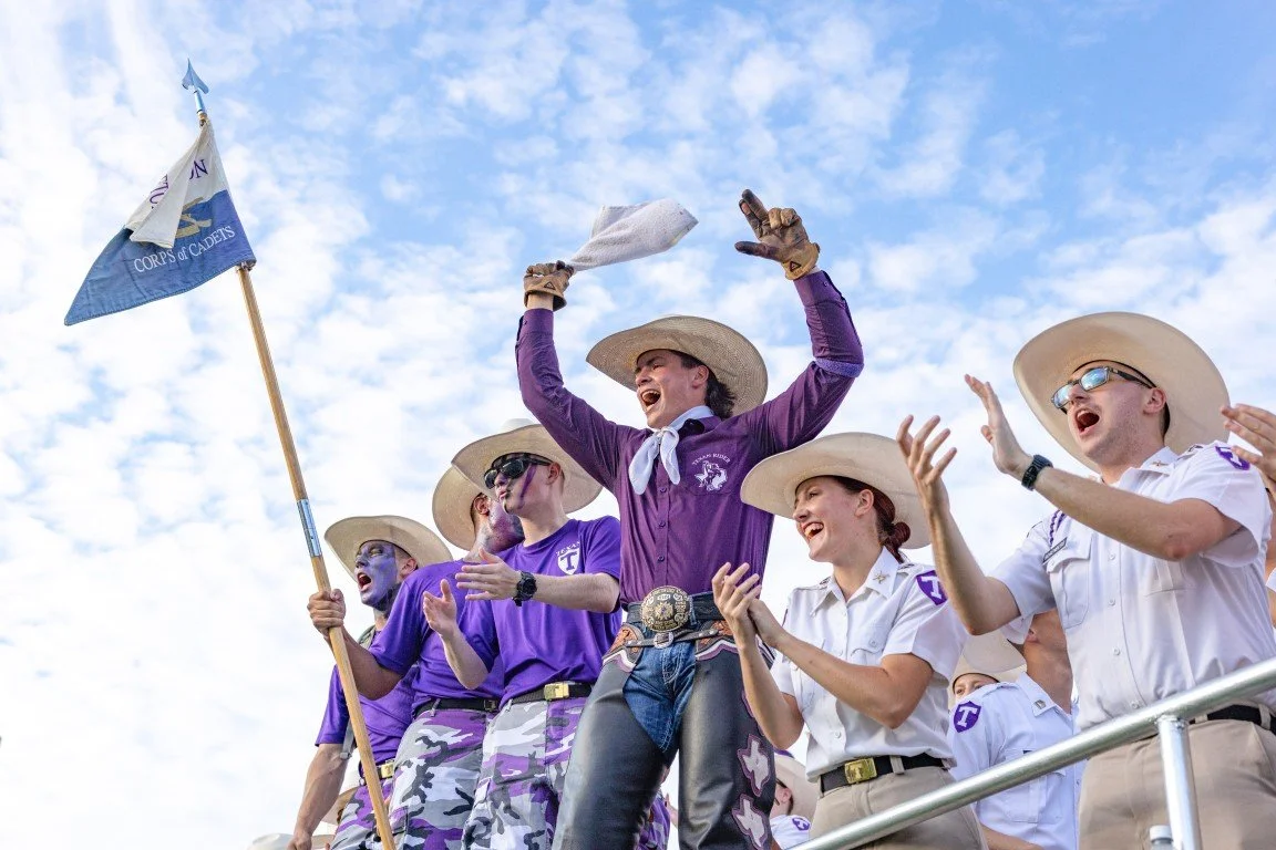 A group of young people celebrating, wearing cowboy hats and purple clothing, with one person holding a flag that says 'Corps of Cadets', against a partly cloudy sky.