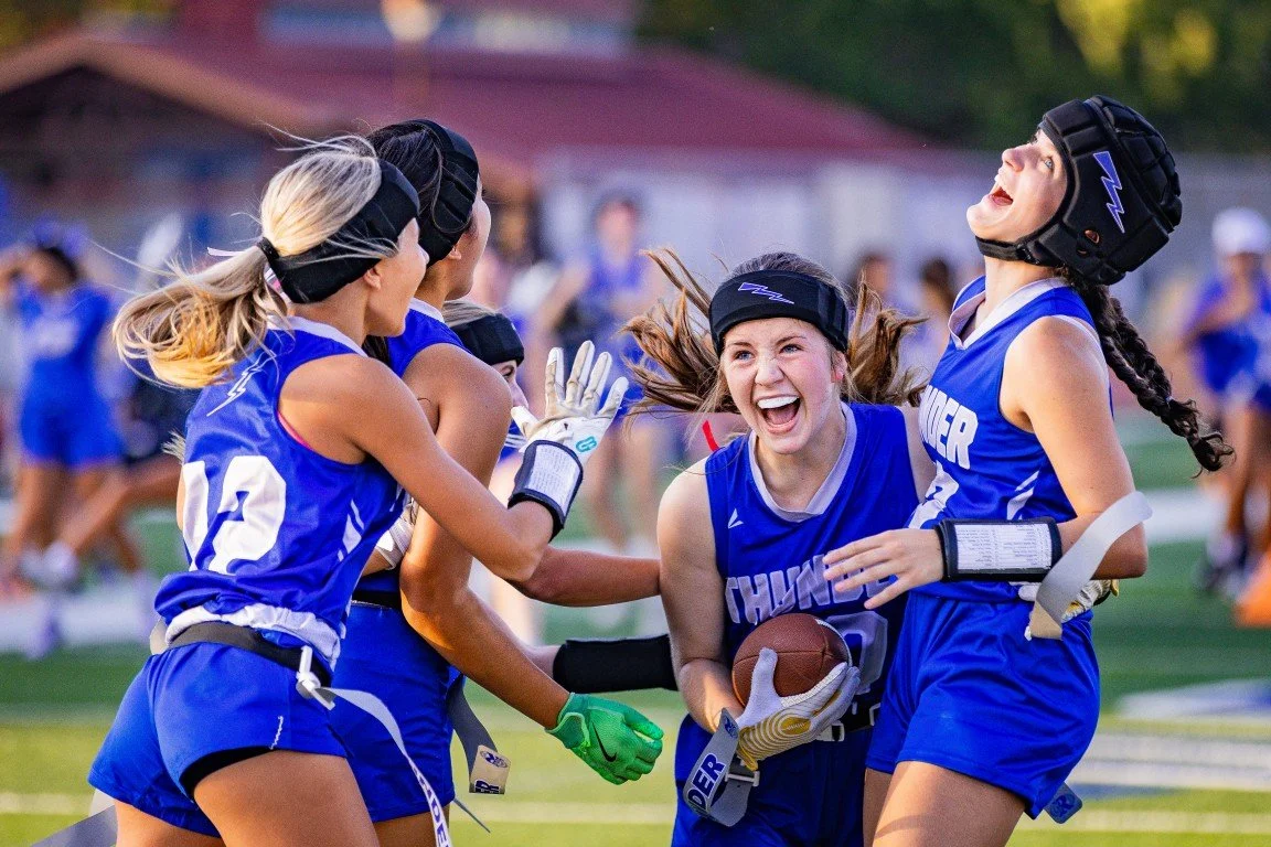 Women’s football players in blue uniforms celebrating on the field after a play, one holding a football, wearing helmets and gloves.