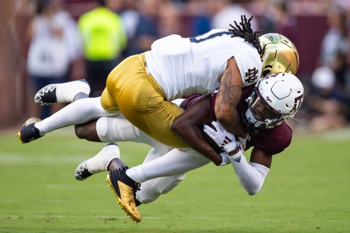 Two football players in action, one in a white and gold uniform tackling the player in a maroon and white uniform on the field with blurred spectators in the background.