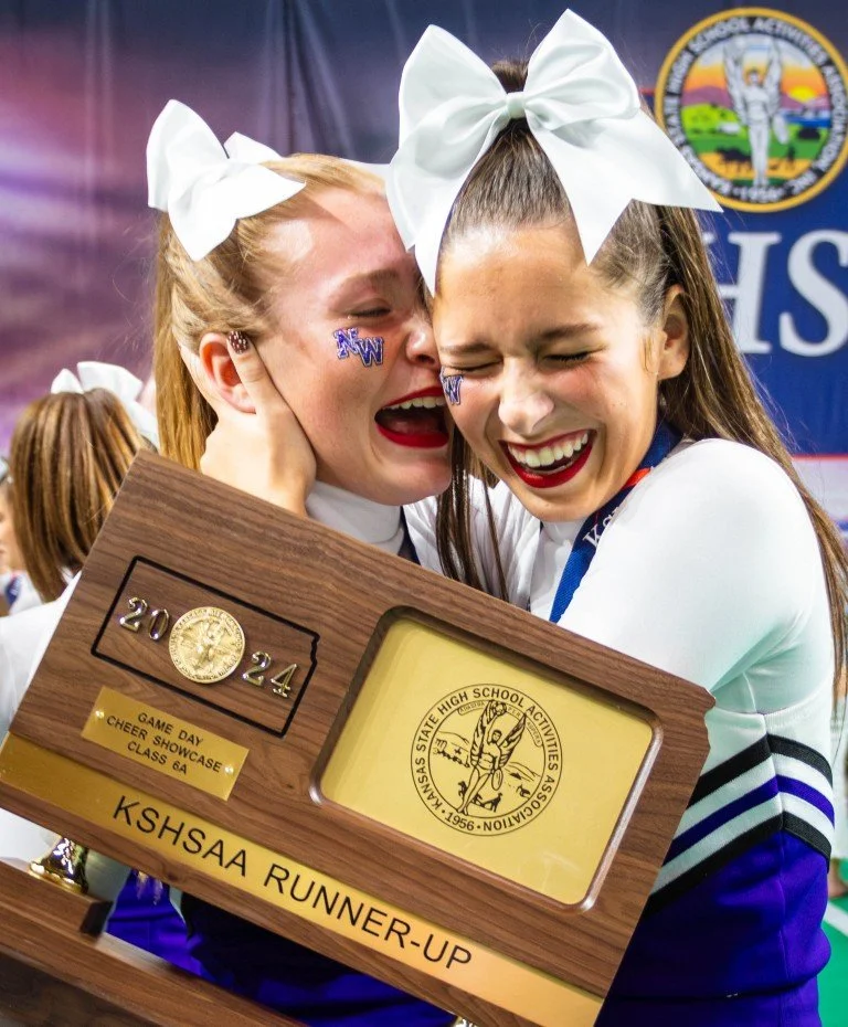 Two cheerleaders celebrating at a high school cheerleading showcase, holding a trophy and a plaque for runner-up in the KSHSAA competition, wearing white bows and cheer uniforms.