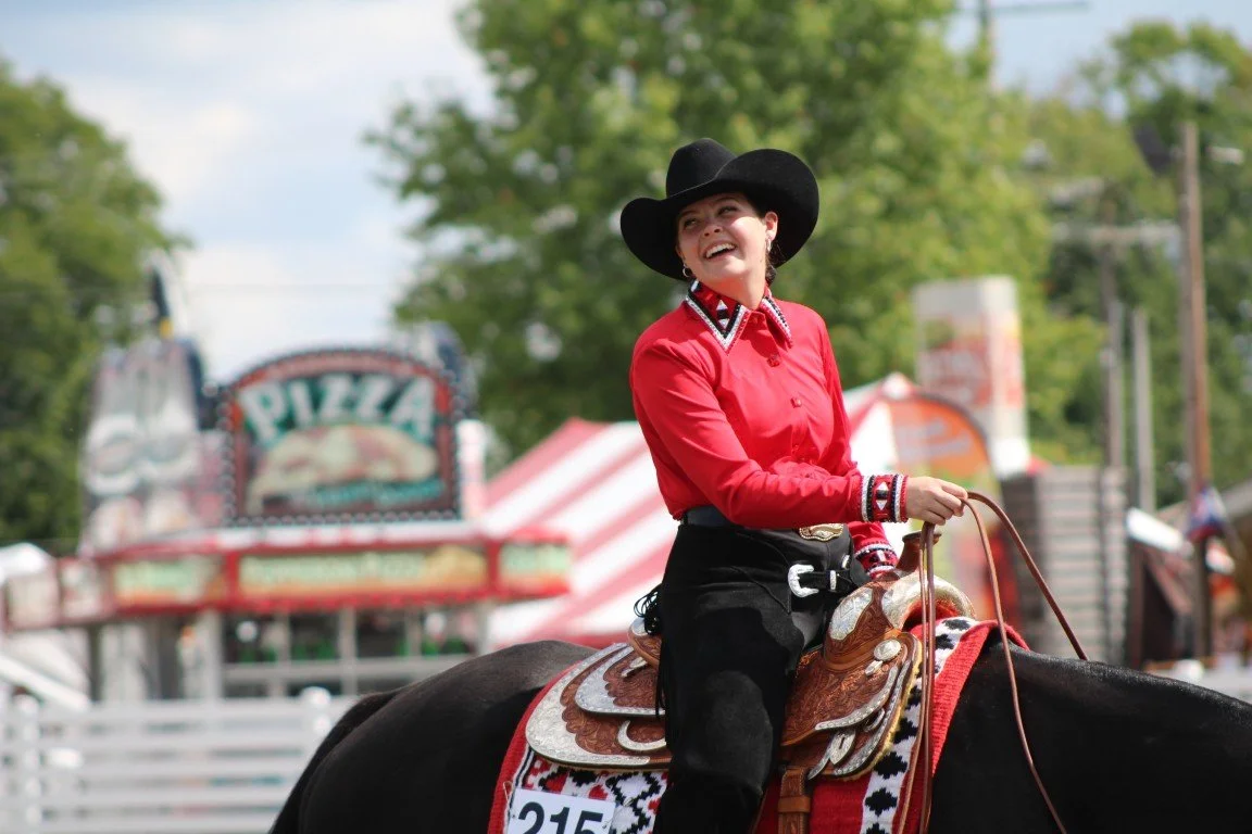 A woman wearing a black cowboy hat, red shirt with decorative collar, and black pants rides a horse at a fair or rodeo. She is smiling and holding the reins, with a colorful horse saddle in front of her. In the background, there are trees and a fairg