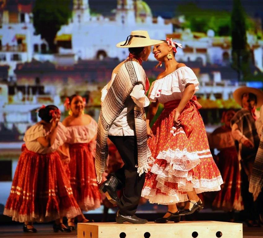 Two dancers on a stage dressed in traditional Mexican attire, performing a folkloric dance, with a colorful background of more dancers in similar costumes.