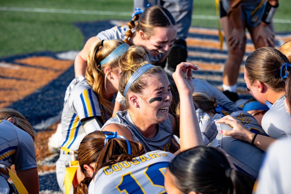 A group of young female softball players in gray and blue uniforms are on the field, with one girl emotionally crying and others comforting her during a game.