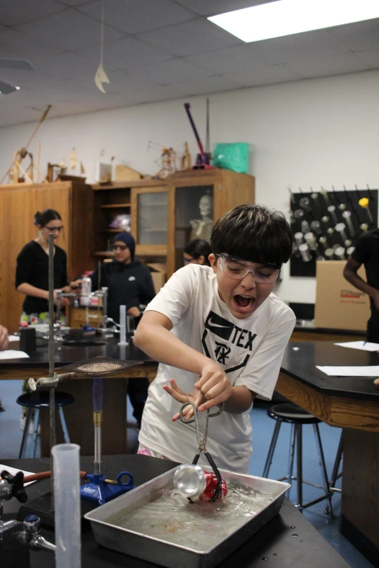 A young boy wearing safety goggles is excitedly working with a science experiment involving a metal tray of water and a musical instrument submerged in it, in a science classroom with other students and lab equipment in the background.