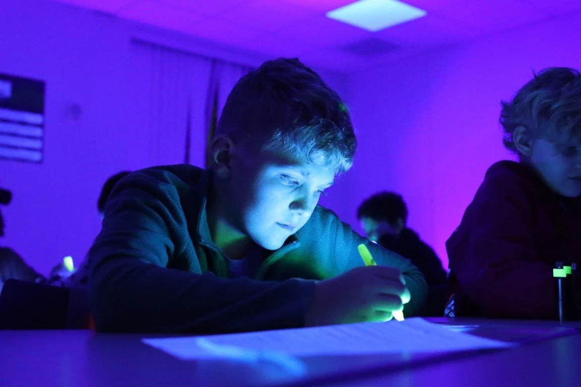 Boy writing with a glow-in-the-dark pen under blacklight in a dark room, other children in the background.