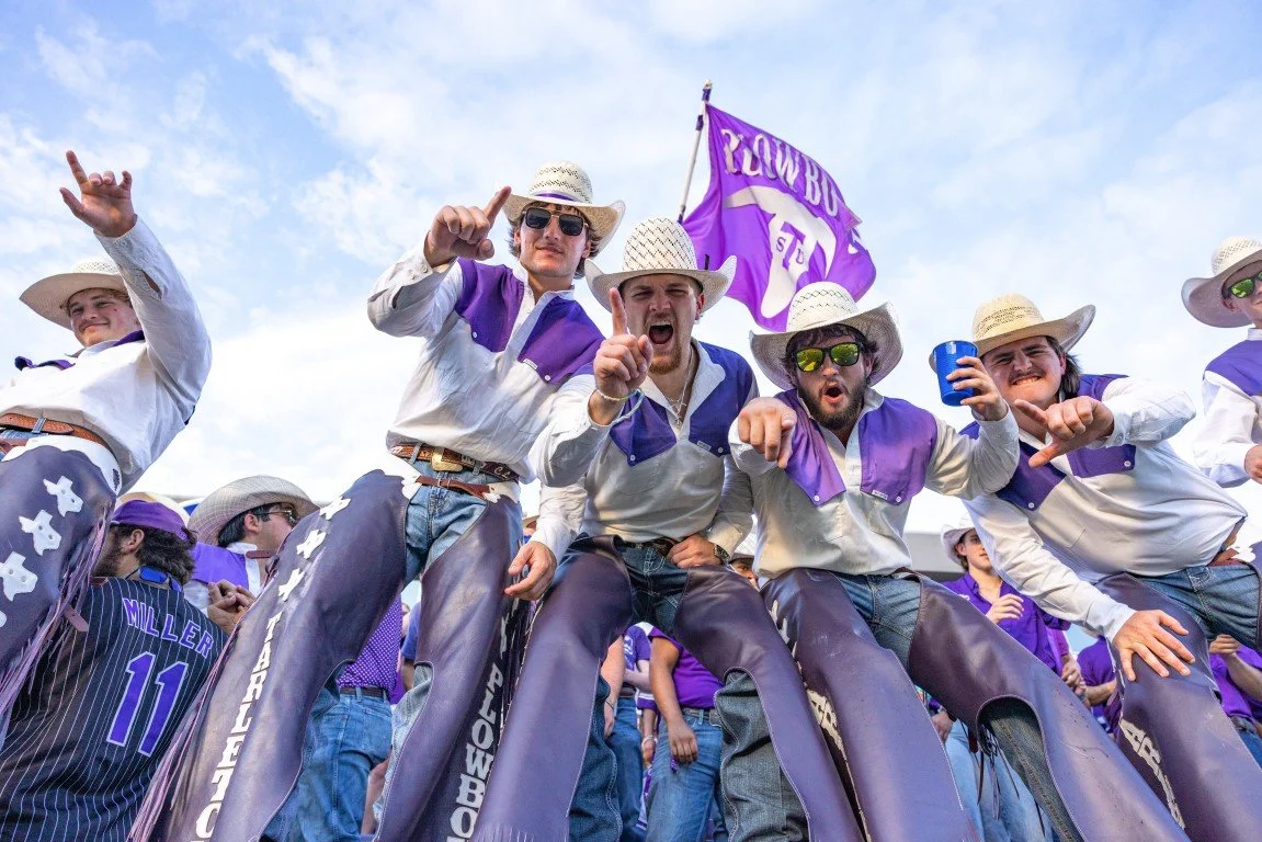 Group of young men wearing straw hats, sunglasses, and purple and white clothing, standing together at a sports event, some making gestures and holding cups, with a purple flag in the background.