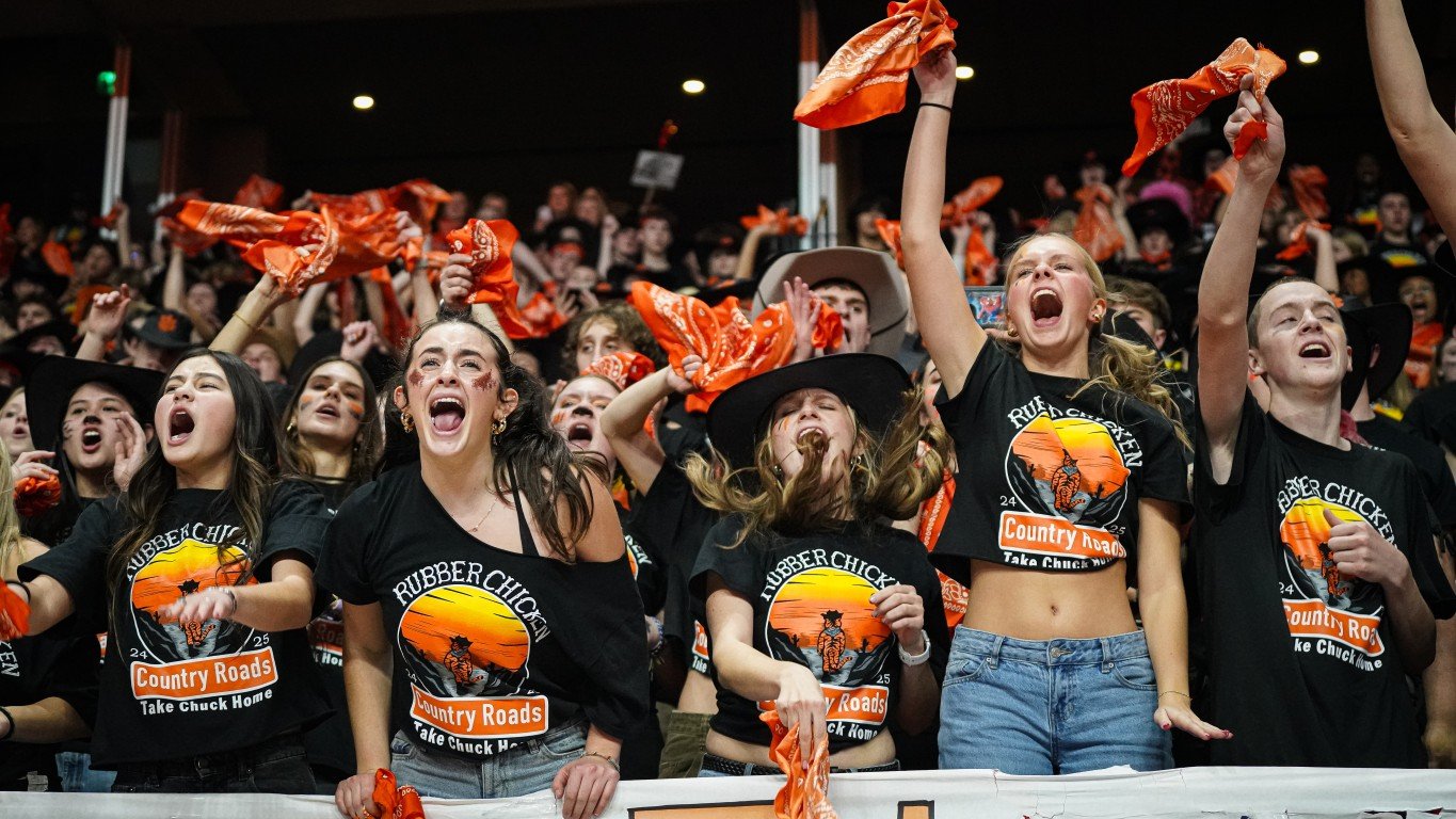 Group of young women cheering and celebrating with orange bandanas and fan signs at a sports event, wearing black T-shirts with a sunset and country theme logo.