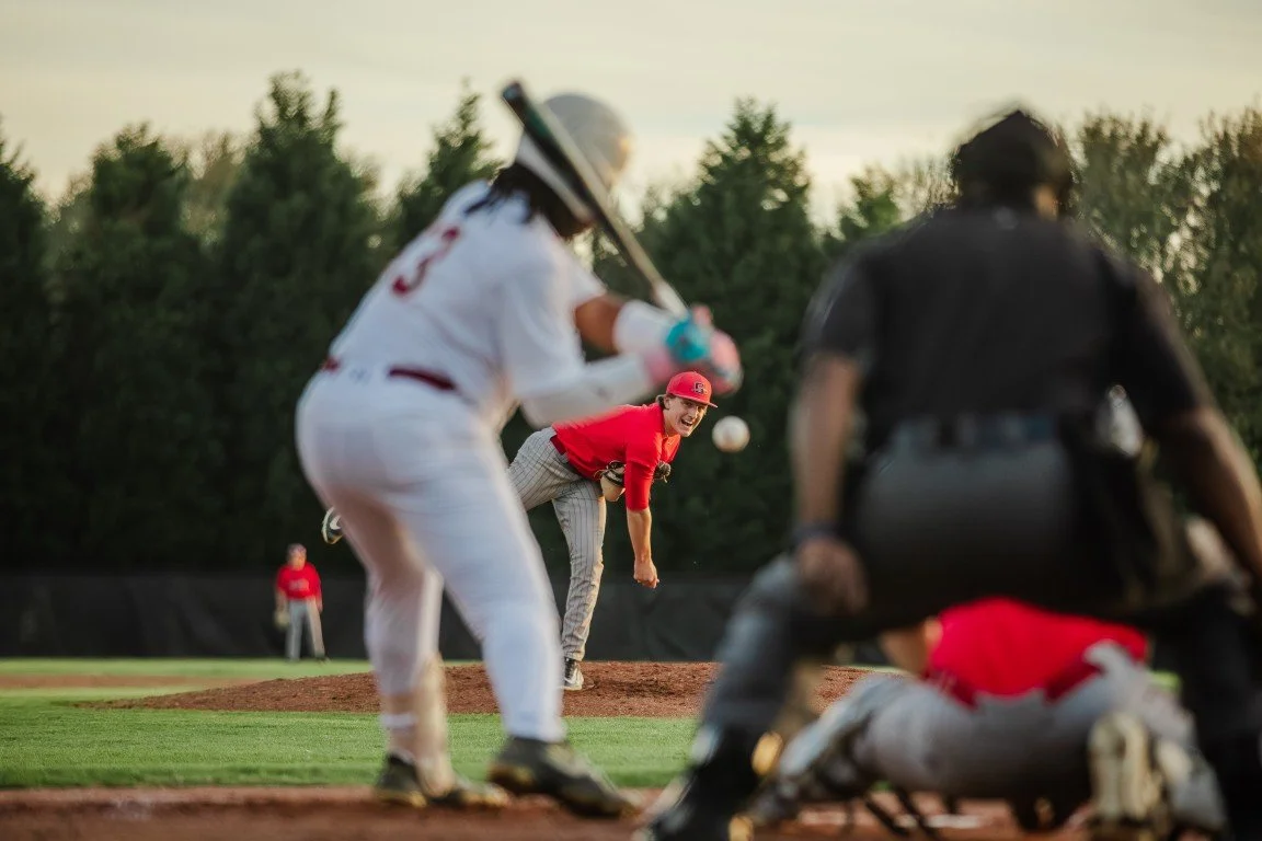 Baseball player in a white uniform ready to catch a pitch, with a pitcher in red and gray mid-throw, and an umpire in black crouched behind the plate, on a baseball field with trees in the background.