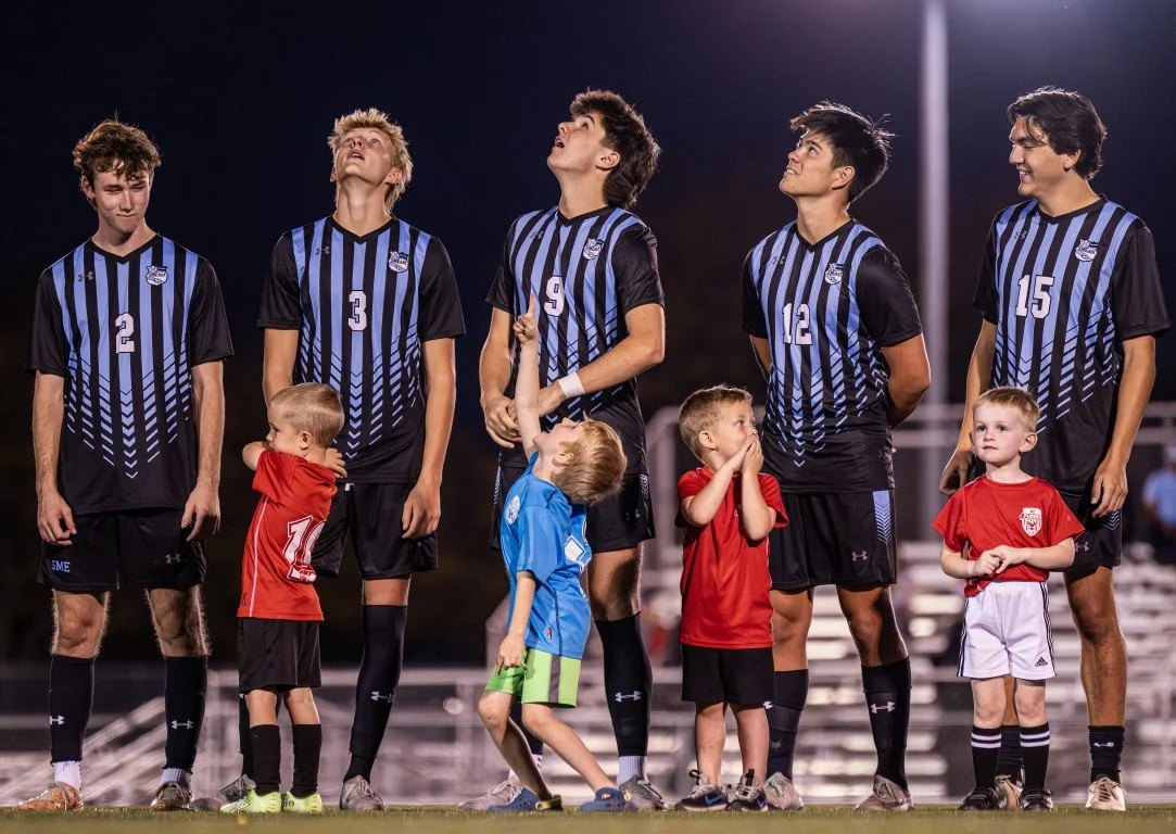 A group of five soccer players in black and blue jerseys and five young children on a soccer field at night. The players are standing in a row, some looking up, with the children in front, some hugging or holding onto the players.