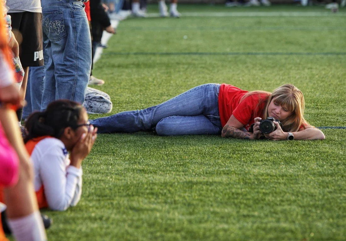 A woman wearing a red shirt and jeans lying on the grass, holding a camera, taking a photo of children at a sports event.
