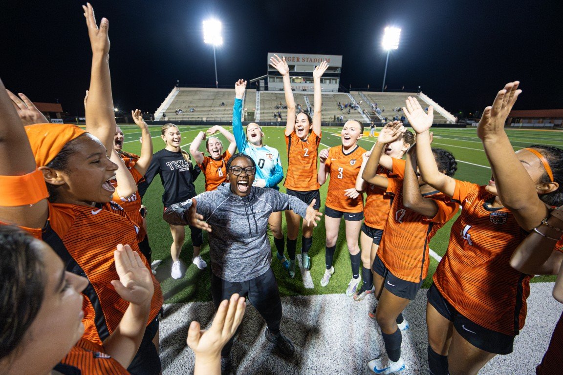 A group of female soccer players and a coach celebrating on a football field at night, with stadium lights illuminating them and empty bleachers in the background.