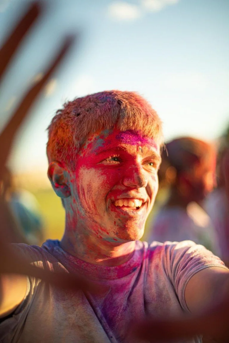 A young person with blush and hair colored pink, smiling and covered in vibrant pink and purple powder during a colorful festival outdoors in daylight.