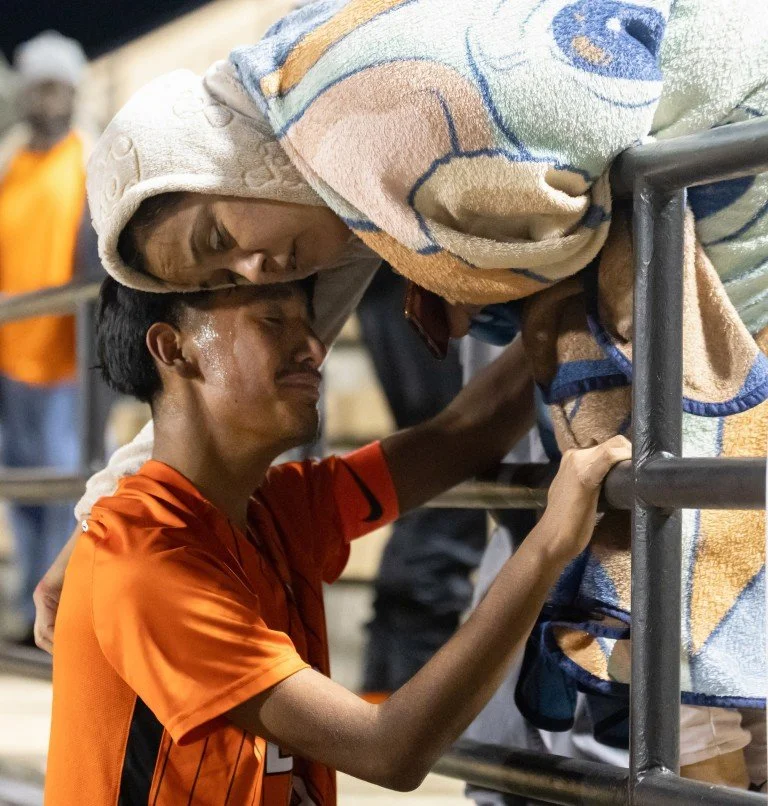 A young man wearing an orange sports jersey leaning over a railing, with a towel on his head, embracing and crying with a woman leaning over the same railing in a sports stadium.