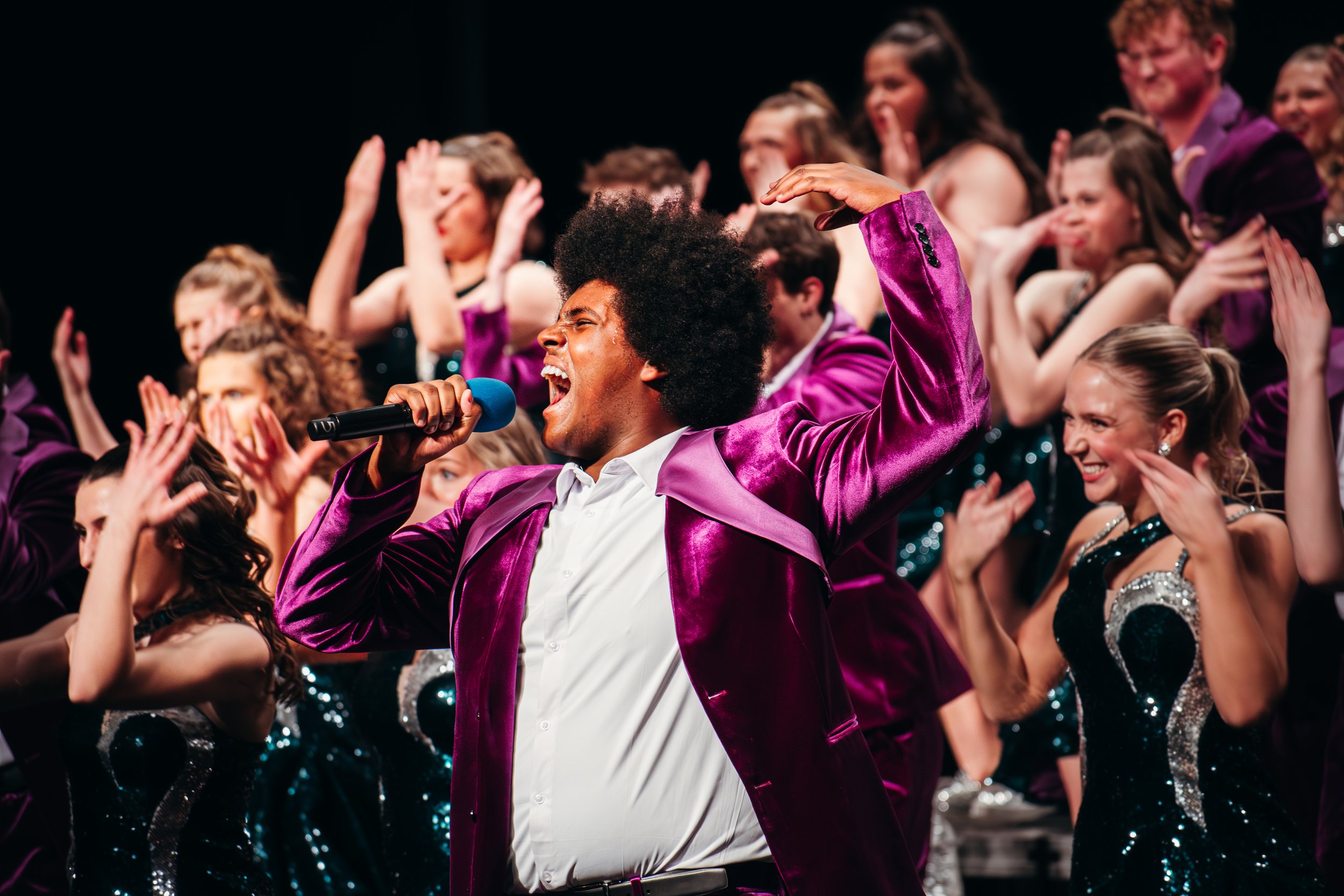 A man with an afro hairstyle singing into a microphone while dancing on stage with a diverse group of people in glamorous outfits.