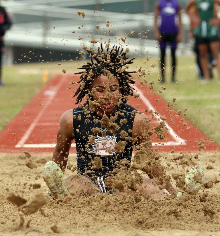 A young male athlete with dreadlocks is jumping into a sandpit during a long jump event, with sand and dirt flying around as he lands.