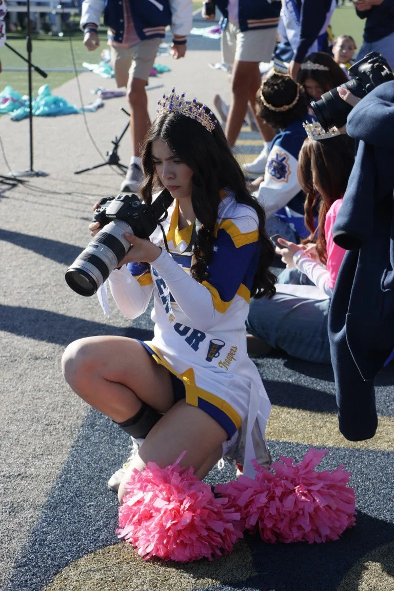 A young woman dressed as a cheerleader, wearing a crown and pom-poms, is kneeling on the ground and looking at a camera she is holding. She is outdoors, surrounded by other people, some of whom are also dressed in cheerleading uniforms, and there are
