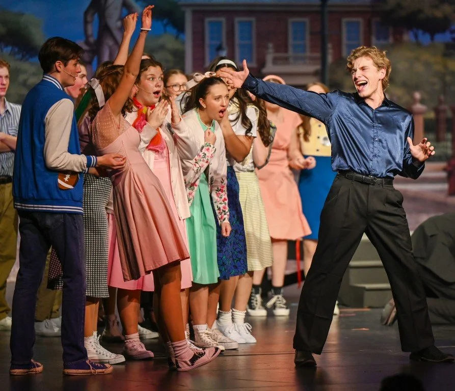 A young man with blonde hair is performing dramatically in front of a group of excited young girls on a stage, with a school or building in the background.