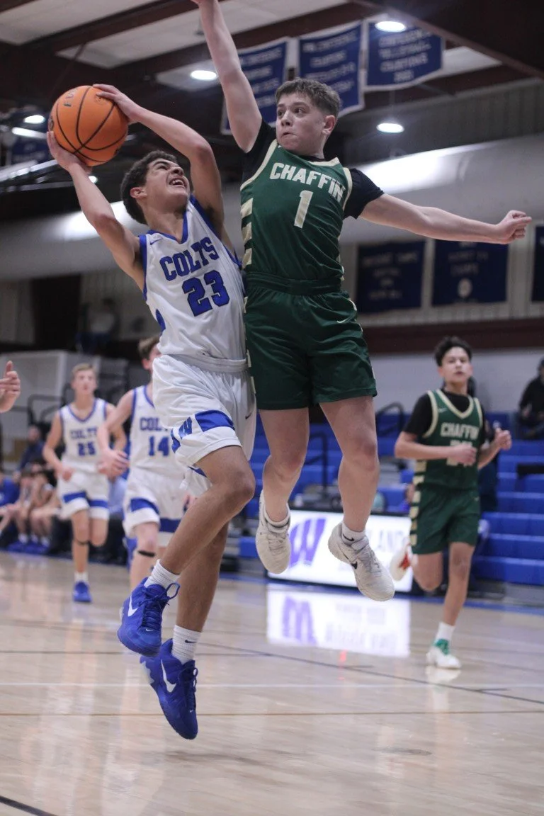 Two young boys playing basketball during a game, one in a white and blue uniform and the other in a green and black uniform, jumping for the ball.