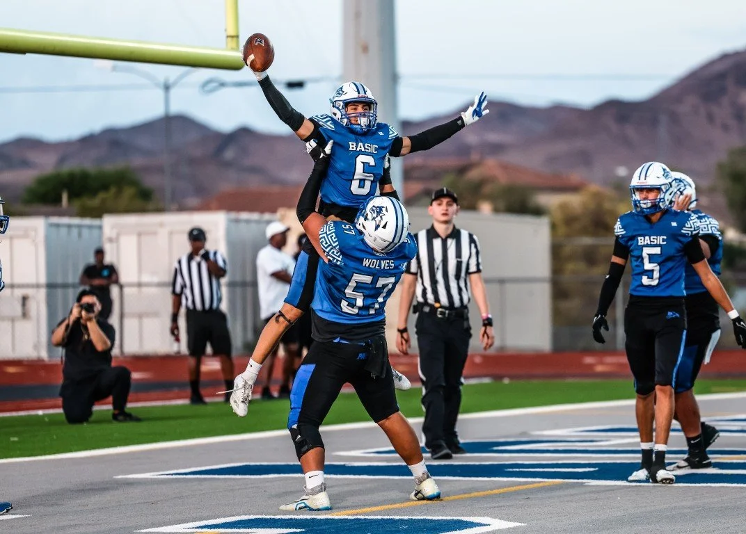 Football players in blue jerseys celebrating a touchdown on the field, with mountains and sky in the background.