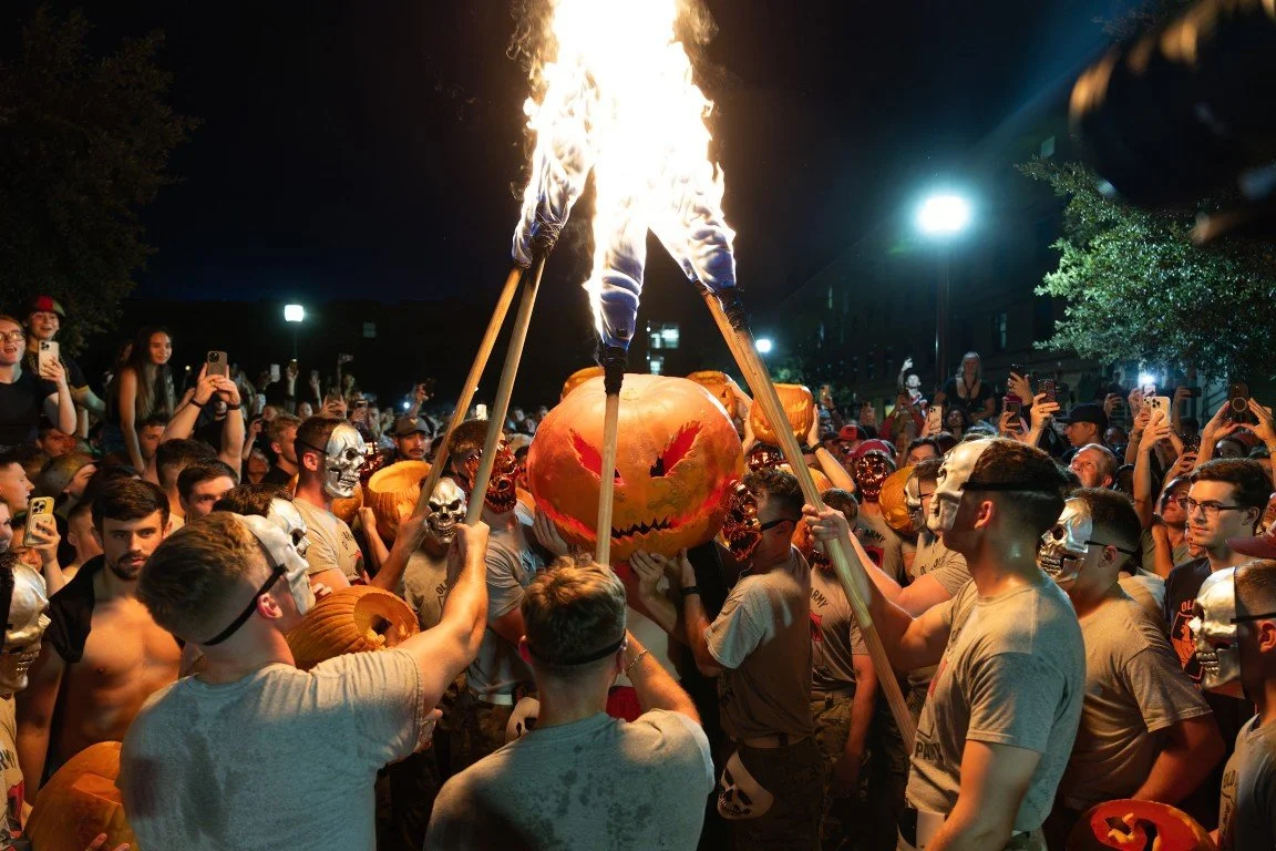 Halloween celebration with people wearing skull masks, holding flames under a carved pumpkin, and taking photos at night.