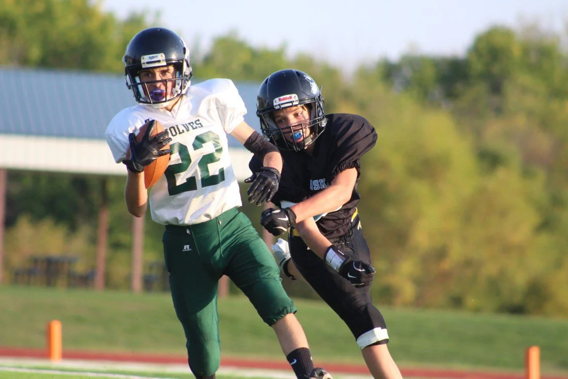 Two youth football players in helmets and uniforms play on a field. One player, in a white jersey with the number 22, carries a football, while the other, in a black jersey, attempts to tackle. Background features a grassy field and trees.