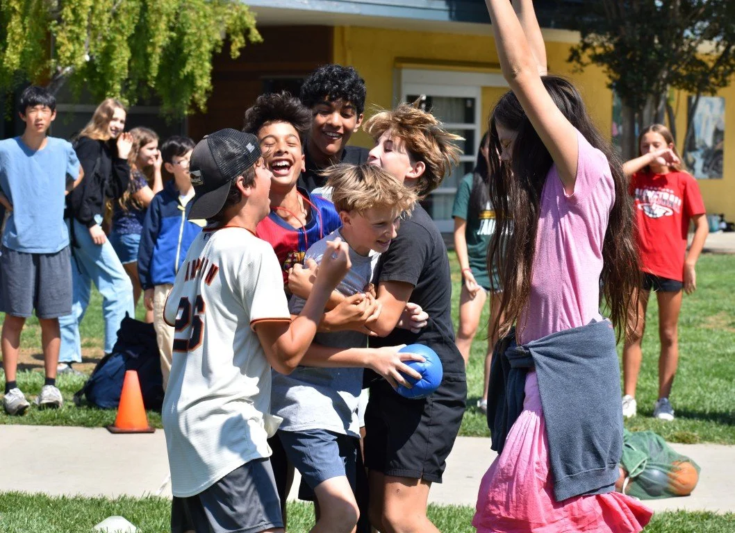 Group of children playing tug of war outdoors on a sunny day, smiling and laughing, with some watching in the background.