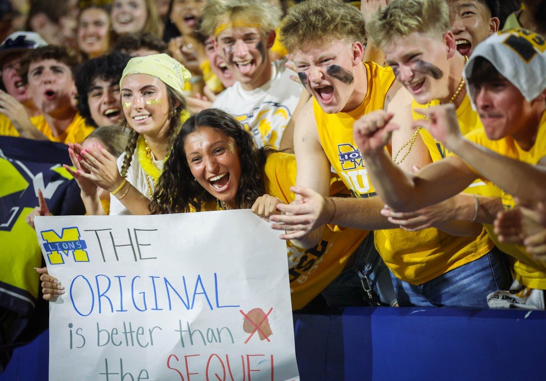 Cheering sports fans, primarily young people, wearing yellow team apparel, holding a sign that reads 'The Original is better than...' in a crowded stadium.